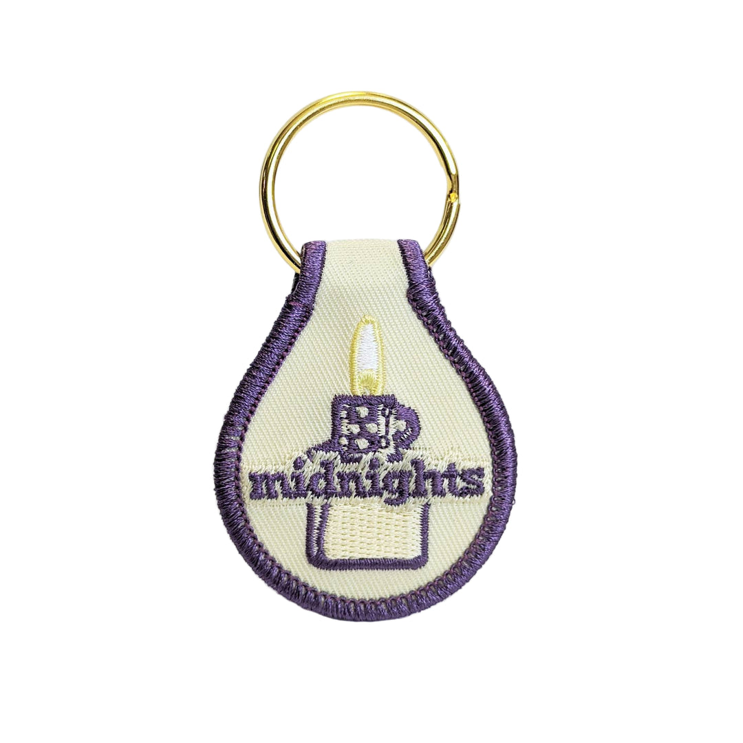 A double-sided fabric key tag with 'Midnights' text embroidered on it, in a purple and beige color scheme, with a key ring.