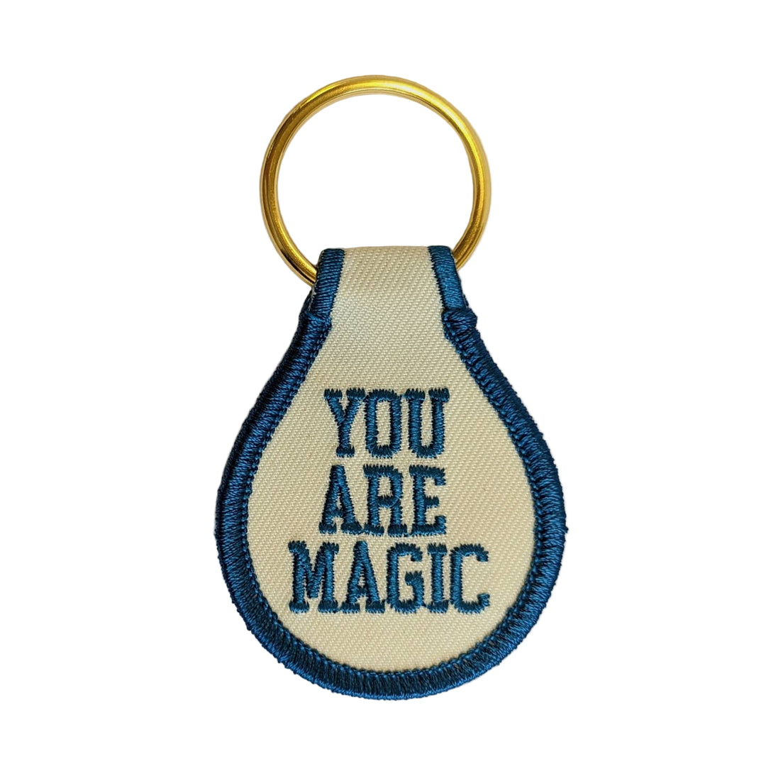 A beige fabric key tag with the text 'YOU ARE MAGIC' embroidered in blue, attached to a gold-colored key ring.
