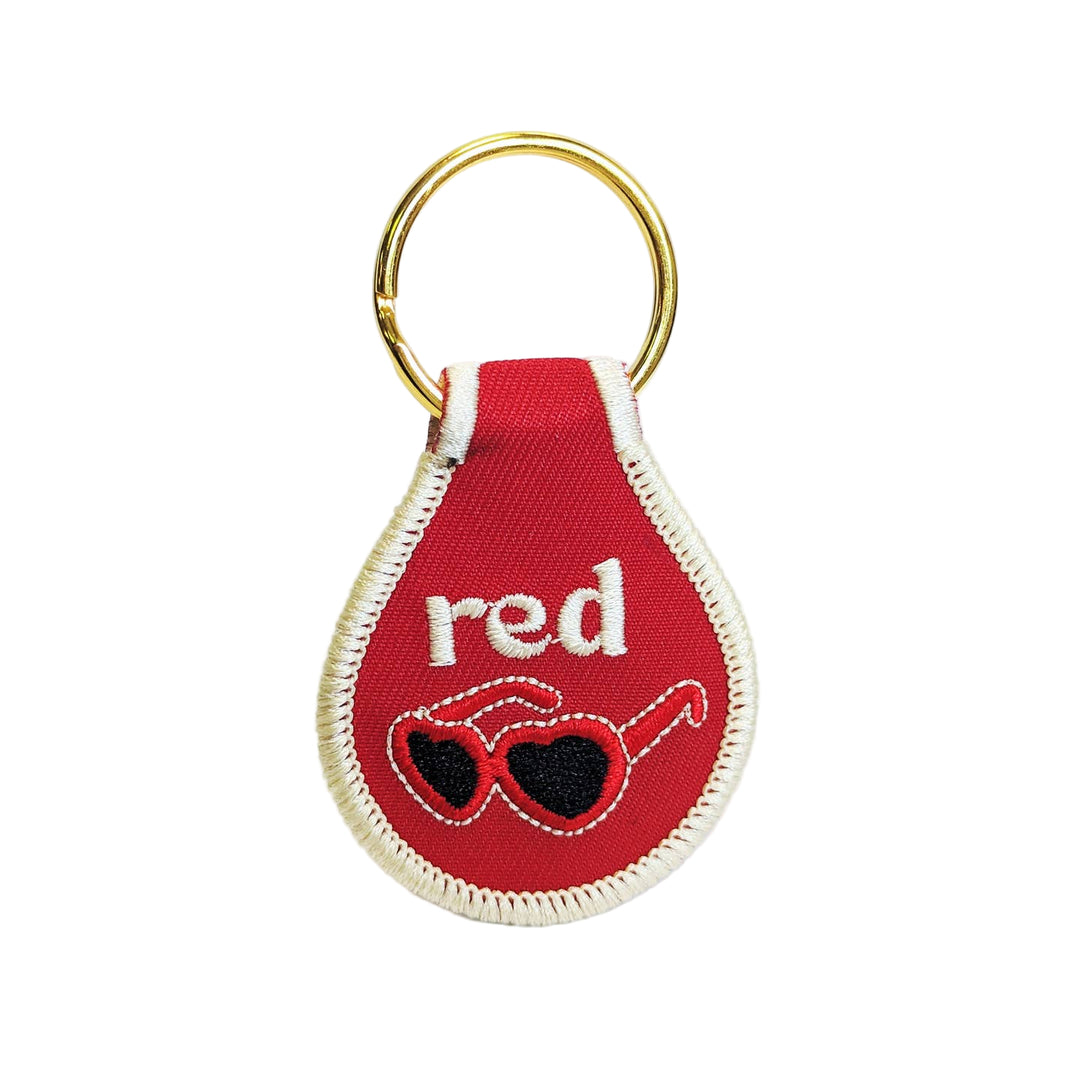 A red fabric key tag with the word 'red' embroidered on it and a pair of sunglasses design, attached to a gold-colored keyring.
