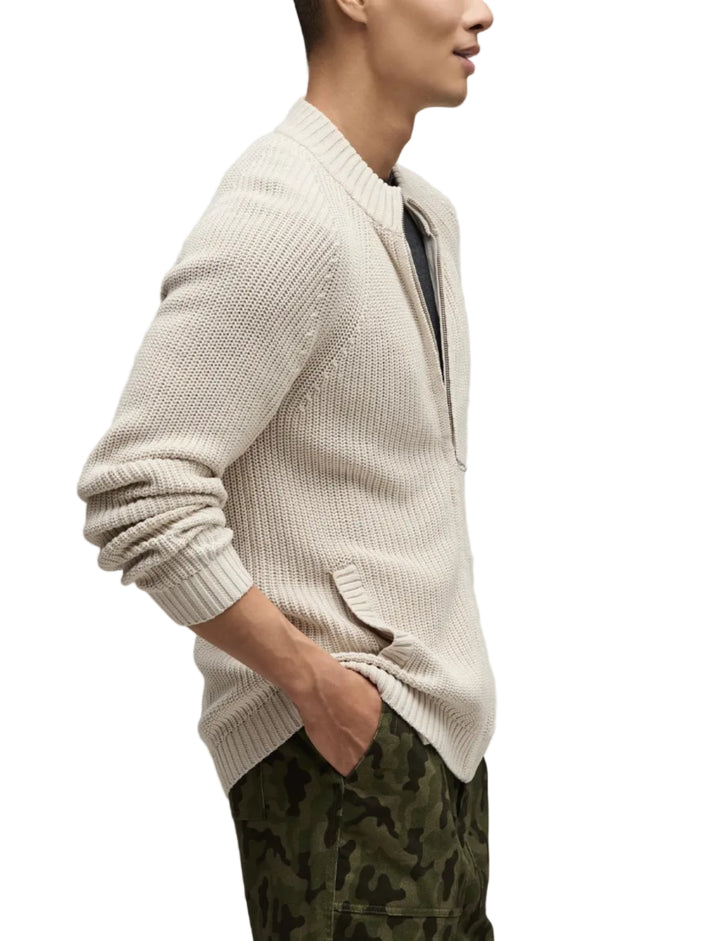 Man wearing a cream cardigan over a dark shirt with camouflage pants on a light gray background