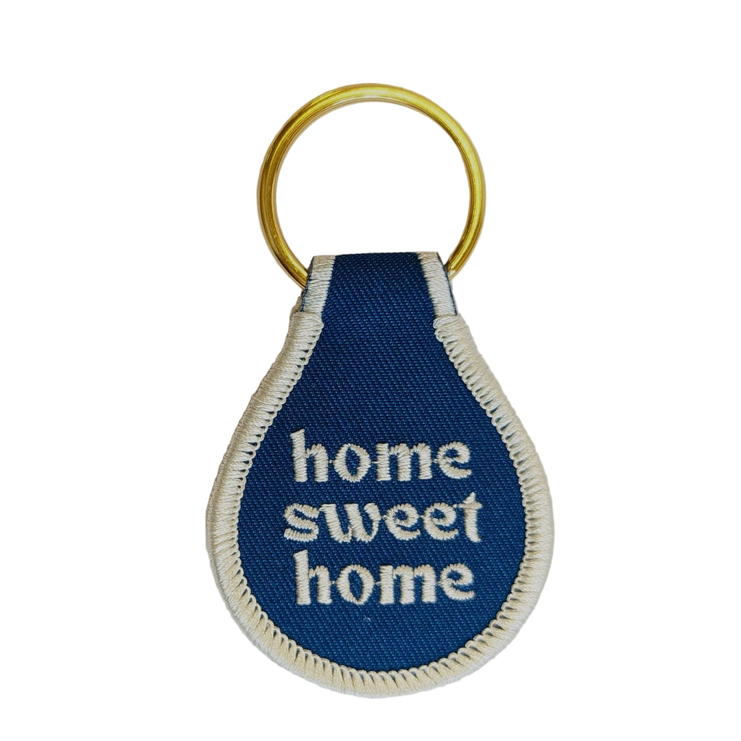 A blue fabric key tag with the text 'home sweet home' embroidered in white, attached to a gold-colored keyring.